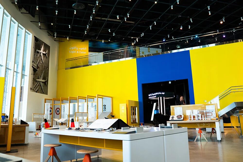 TSCK gallery with bright yellow and blue walls, exhibit tables and a black ceiling with rows of track spotlights