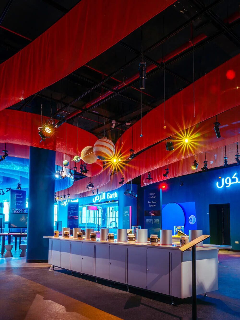Gallery with red fabric ceiling, suspended planet models and a long white exhibit counter under track spotlights