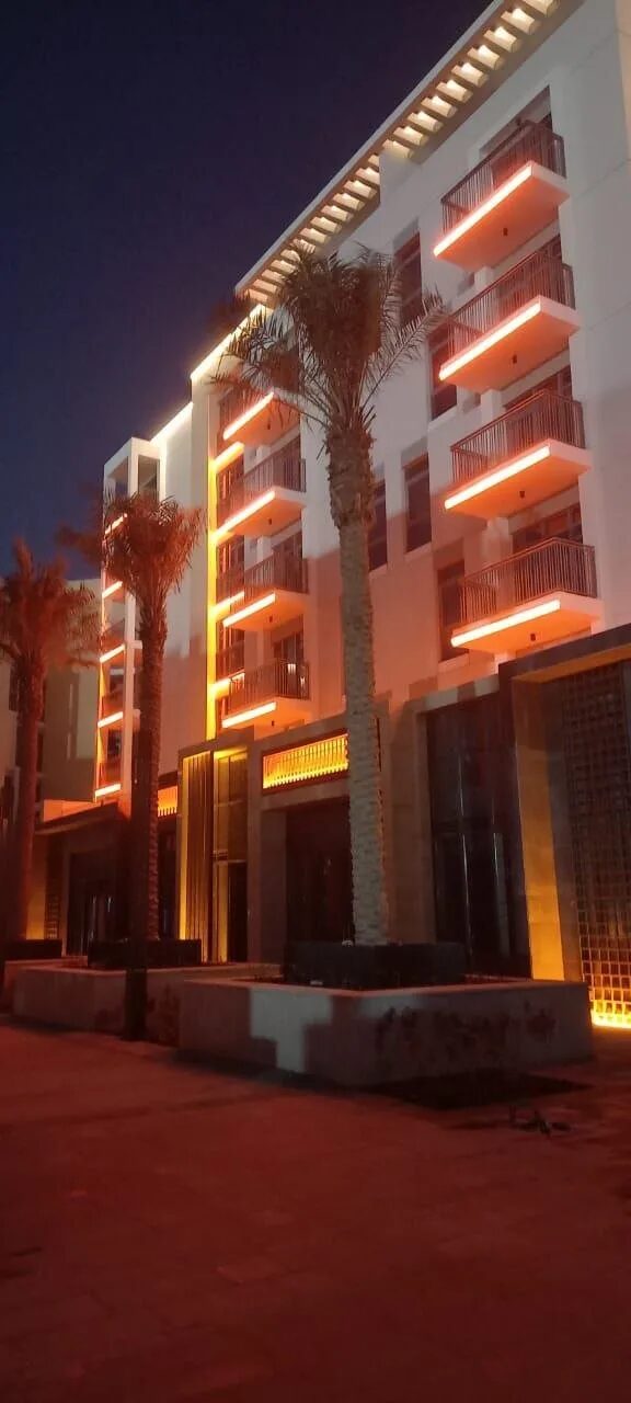Tall residential block at night with amber light lines under each balcony and palm trees in front