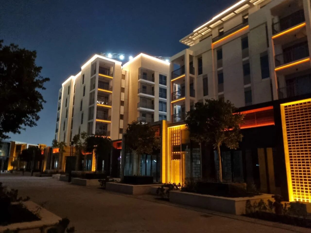 Street at Al Zahia Uptown at night with residential blocks, warm linear light along balconies and lit lattice screens at ground level